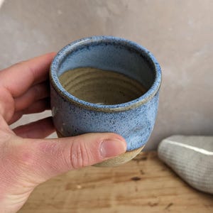May include: A handmade ceramic cup with a light blue exterior and a tan interior. The cup has a slightly textured surface and a rounded shape. The rim is a darker shade of blue. The cup is held in a hand.