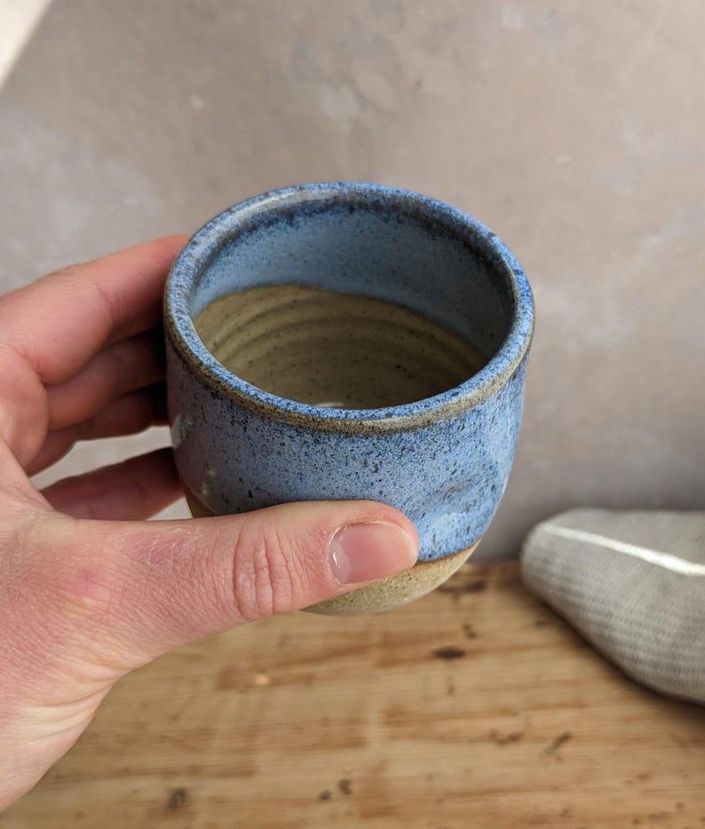 May include: A handmade ceramic cup with a light blue exterior and a tan interior. The cup has a slightly textured surface and a rounded shape. The rim is a darker shade of blue. The cup is held in a hand.