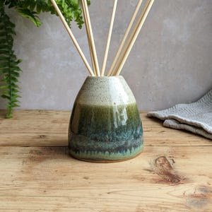 May include: A ceramic reed diffuser vessel with a tapered shape, featuring a speckled cream top that transitions into a deep forest green and teal base. Several light-colored wooden reeds are inserted into the opening of the container.