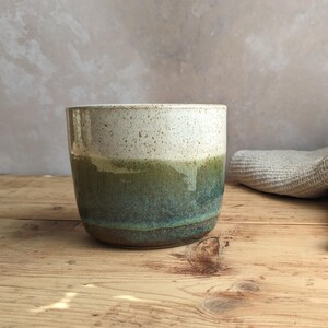 May include: A handmade ceramic mug with a speckled white top, transitioning to a green and teal glazed body. The mug sits on a wooden surface, showcasing its unique, handcrafted design. The mug is approximately 4 inches tall.
