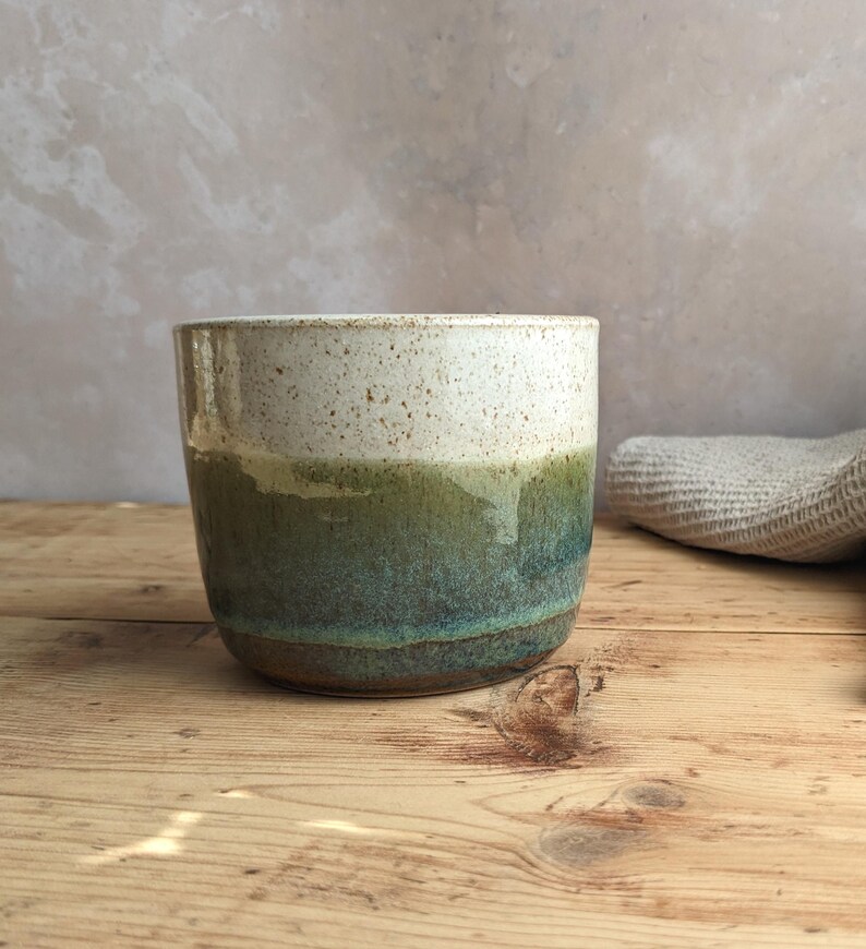 May include: A handmade ceramic mug with a speckled white top, transitioning to a green and teal glazed body. The mug sits on a wooden surface, showcasing its unique, handcrafted design. The mug is approximately 4 inches tall.