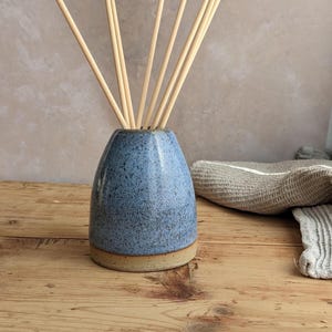 May include: A blue ceramic essential oil diffuser with a speckled finish and a natural base, filled with wooden reeds. The diffuser sits on a wooden surface, with a neutral-toned background. A folded cloth is in the background.