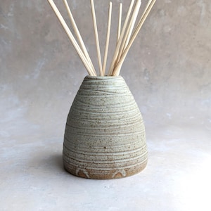 Könnte beinhalten: Ein Keramik-Duftdiffusor in hellem Grau mit strukturierter Oberfläche. Der vasenförmige Behälter enthält mehrere hellfarbene Holzstäbchen, die eine natürliche Ästhetik erzeugen. Der Diffusor ist für Raumduft konzipiert.