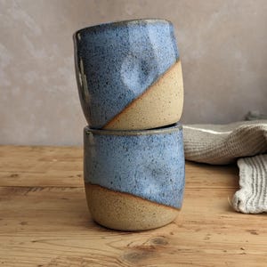 May include: Two handmade ceramic cups stacked on top of each other. The cups are a light blue color on the top half and a tan color on the bottom half, with a diagonal color split. The cups are sitting on a wooden surface.