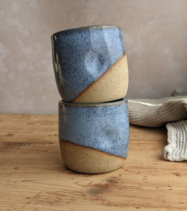 May include: Two handmade ceramic cups stacked on top of each other. The cups are a light blue color on the top half and a tan color on the bottom half, with a diagonal color split. The cups are sitting on a wooden surface.