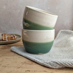 May include: Two stacked ceramic bowls featuring a speckled off-white upper section and a solid deep green lower section. The bowls have a smooth, rounded silhouette with a wide opening and a tapered base, showcasing a two-tone glazed finish.