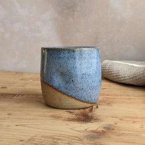 May include: A handmade ceramic cup with a speckled blue glaze on the upper portion and a natural tan base. The cup has a rounded shape and is set on a wooden surface. The background is a neutral color.