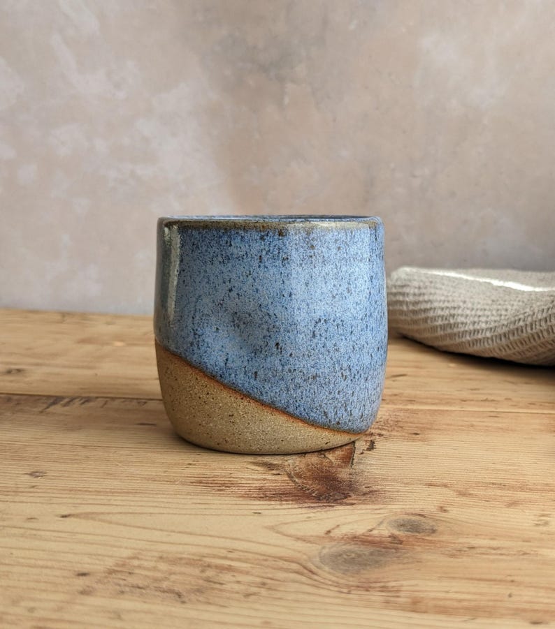 May include: A handmade ceramic cup with a speckled blue glaze on the upper portion and a natural tan base. The cup has a rounded shape and is set on a wooden surface. The background is a neutral color.