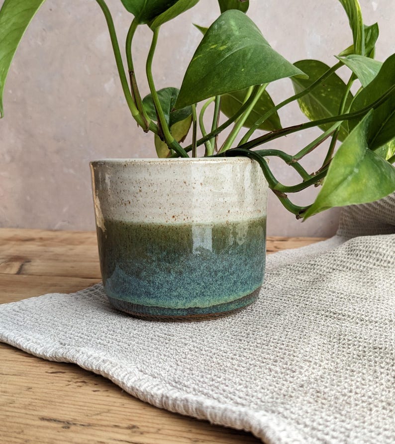 May include: A ceramic plant pot with a two-tone design. The top half is a speckled off-white, transitioning to a deep green and then a teal blue at the base. The pot holds a green leafy plant. The pot sits on a light gray textured cloth.