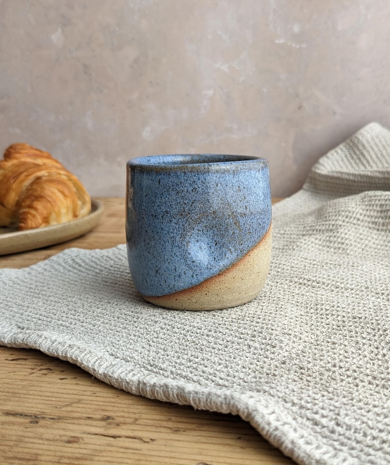 May include: A handmade ceramic mug with a blue and tan glaze. The mug has a rounded shape and a smooth finish. A croissant sits on a plate in the background. The mug is on a gray linen napkin on a wooden table.