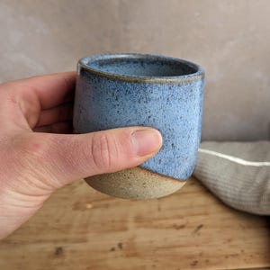 May include: A handmade ceramic tumbler with a speckled blue glaze and a natural, unglazed base. The cup is held in a hand, showcasing its rounded shape and rustic aesthetic. The background features a wooden surface and a neutral-toned backdrop.