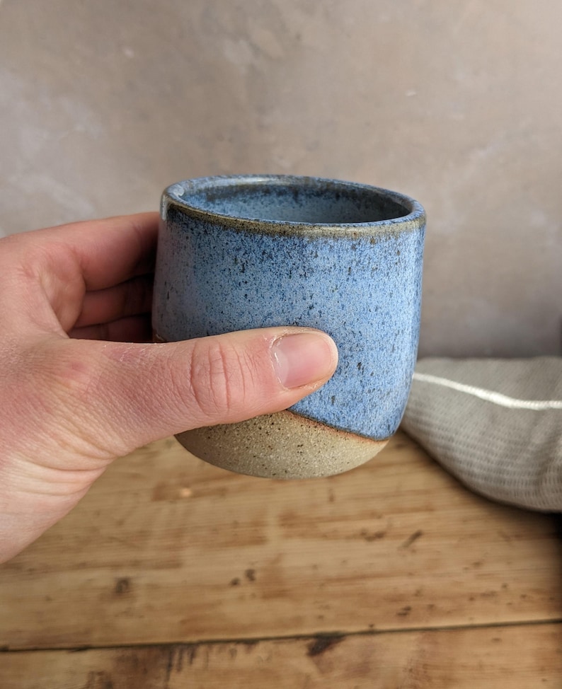 May include: A handmade ceramic tumbler with a speckled blue glaze and a natural, unglazed base. The cup is held in a hand, showcasing its rounded shape and rustic aesthetic. The background features a wooden surface and a neutral-toned backdrop.