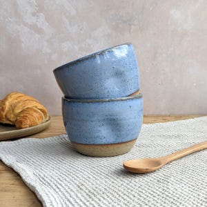 May include: Two stacked ceramic bowls with a speckled light blue glaze and a natural tan base. The bowls feature a slightly irregular, hand-formed shape with subtle indentations along the sides, showcasing a textured finish and earthy tones.