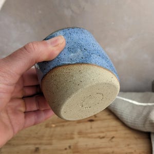 May include: A handmade ceramic tumbler with a speckled blue glaze on the upper portion and a natural, unglazed tan base. The cup has a rounded shape and a smooth, tactile finish, perfect for enjoying beverages.