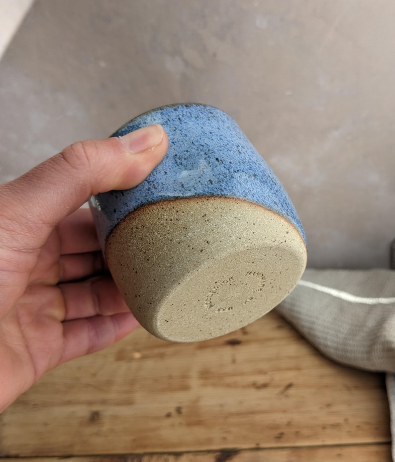 May include: A handmade ceramic tumbler with a speckled blue glaze on the upper portion and a natural, unglazed tan base. The cup has a rounded shape and a smooth, tactile finish, perfect for enjoying beverages.