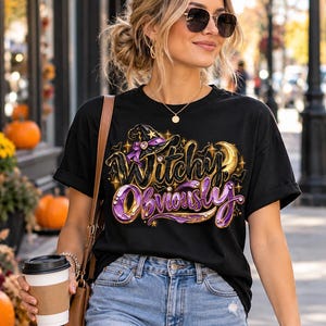 May include: A black short-sleeved t-shirt featuring a graphic print with the text Witchy Obviously in purple and gold lettering. The design includes a black witch hat with a purple ribbon, a gold crescent moon, and small star accents.