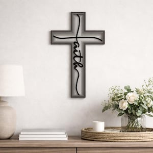May include: A layered, gray wooden cross with the word "faith" in a cursive font. The cross is mounted on a white wall. A lamp, books, and a floral arrangement are on a wooden table below.