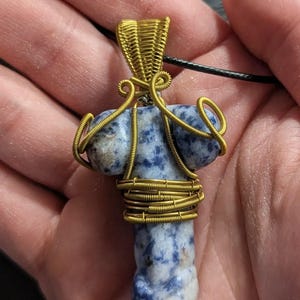 Könnte beinhalten: Ein Anhänger mit einem blau-weißen Stein, der kunstvoll mit goldfarbenem Draht umwickelt ist. Der Anhänger hängt an einer schwarzen Kordel. Der Stein hat eine einzigartige Form, und die Drahtarbeit verleiht dem Schmuck ein dekoratives Element.