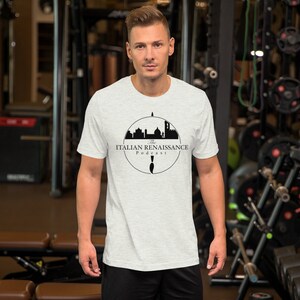May include: A light gray t-shirt featuring the text "The Italian Renaissance Podcast" in black, along with a silhouette of Italian architecture. The design is enclosed in a circular frame. The shirt is short-sleeved.