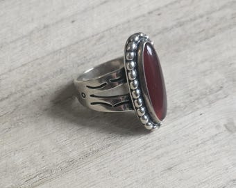 Könnte beinhalten: Ein silberner Ring mit einem ovalen, tiefroten Stein. Der Stein ist von einer perlenbesetzten Bordüre umgeben. Das Ringband hat dekorative Ausschnitte. Der Ring liegt auf einer hellen Holzmaserung.