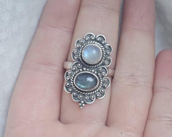 May include: A silver ring featuring two oval-shaped gemstones. The top stone is a white moonstone, and the bottom stone is a dark grey labradorite. The ring has a floral design.