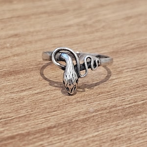 May include: A silver-toned metal ring featuring a coiled snake design. The snake's head is positioned at the centre, with its body wrapping around the band. The metal has a polished finish with darkened recessed areas to highlight the snake's scales.