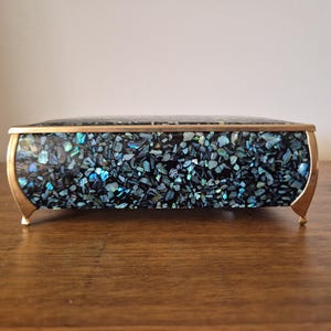May include: A rectangular box featuring an exterior surface covered in a mosaic of iridescent abalone shell fragments in shades of blue, green, and black. The box is framed with a gold-toned metal trim and stands on four small, curved metal feet.