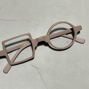 Op de afbeelding: Unieke bril met een modern design. De montuur is een gedempte mauve kleur, met één vierkante lens en één ronde lens. De pootjes en de brug zijn ook mauve. De bril staat op een neutrale achtergrond.