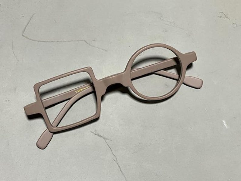 May include: Unique eyeglasses with a modern design. The frames are a muted mauve color, featuring one square lens and one circular lens. The arms and bridge are also mauve. The glasses are set against a neutral background.