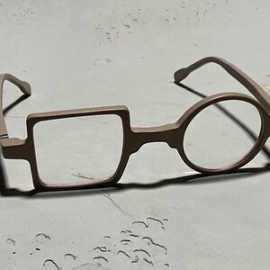 May include: Unique eyeglasses with a square lens on the left and a round lens on the right. The frames are a muted rose color, and the glasses are set against a neutral background.