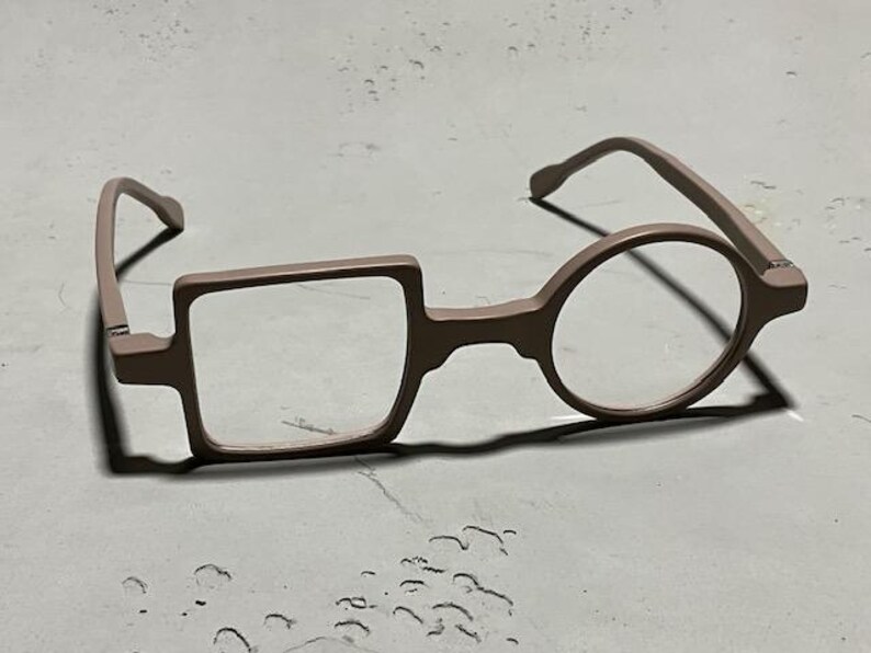 May include: Unique eyeglasses with a square lens on the left and a round lens on the right. The frames are a muted rose color, and the glasses are set against a neutral background.
