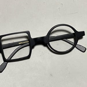Może przedstawiać: Czarne okulary o unikalnym designie. Jedna soczewka jest kwadratowa, druga okrągła. Oprawka jest czarna z napisem "WWW.monkeyeye" na kwadratowej oprawce soczewki. Okulary leżą na jasnoszarej powierzchni.