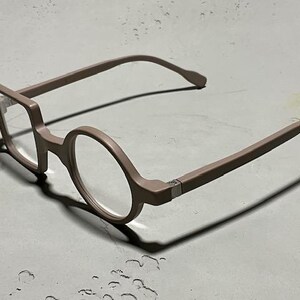 May include: A pair of eyeglasses with a unique design. The frames are a muted mauve color, with one lens in a square shape and the other in a circle. The temples curve gently, and the glasses cast a shadow on the surface.