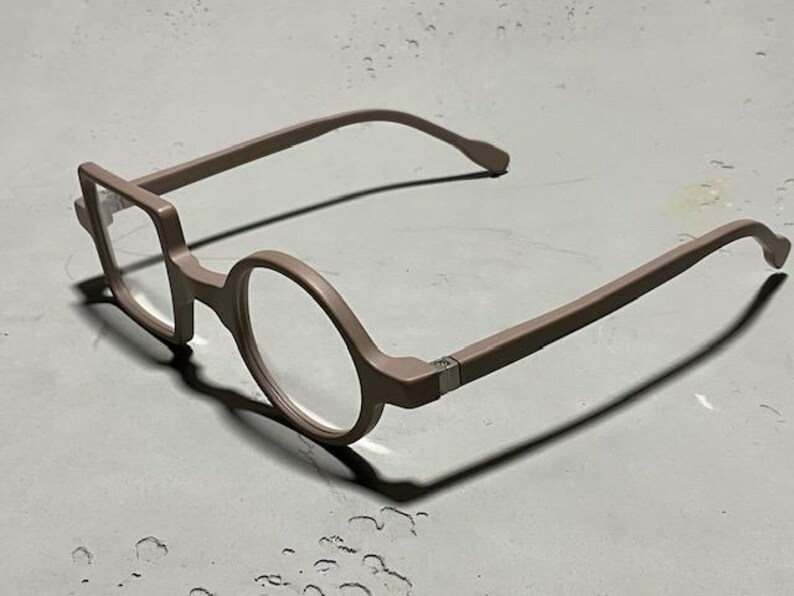 May include: A pair of eyeglasses with a unique design. The frames are a muted mauve color, with one lens in a square shape and the other in a circle. The temples curve gently, and the glasses cast a shadow on the surface.