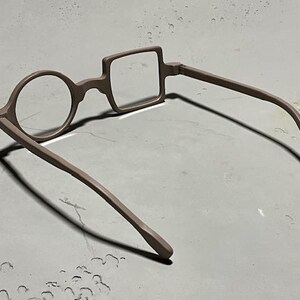 May include: Unique eyeglasses with a round lens on one side and a square lens on the other. The frames and arms are a muted brown color. The glasses are designed with a distinctive, artistic style.