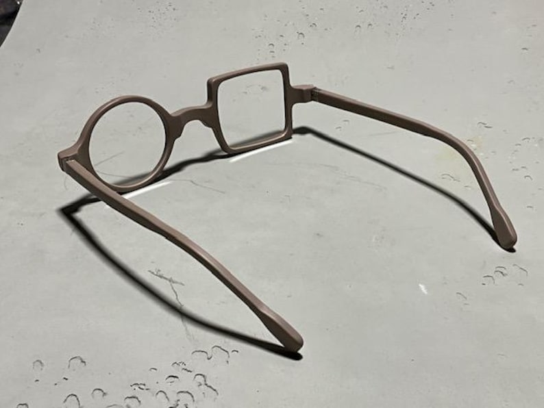 May include: Unique eyeglasses with a round lens on one side and a square lens on the other. The frames and arms are a muted brown color. The glasses are designed with a distinctive, artistic style.