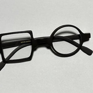 Może przedstawiać: Czarne okulary o unikalnym designie. Jedna soczewka jest kwadratowa, druga okrągła. Oprawka ma elegancki, nowoczesny wygląd. Tekst "www.monkey" jest widoczny na oprawce. Okulary są na gładkim tle.