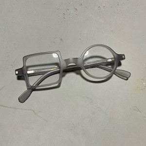 Może przedstawiać: Szare okulary o unikalnym designie. Jedna soczewka jest kwadratowa, druga okrągła. Oprawki i zauszniki są w pasującym szarym kolorze. Okulary leżą na jasnoszarej powierzchni.