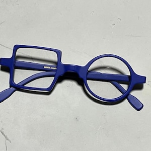 Może przedstawiać: Niebieskie okulary o unikalnym designie. Jedna soczewka jest kwadratowa, druga okrągła. Oprawka jest w jednolitym niebieskim kolorze. Na oprawce wydrukowano tekst "www.monk".
