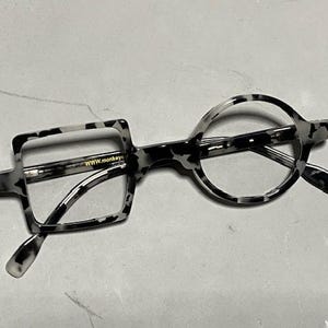 Może przedstawiać: Unikalne okulary z kwadratową soczewką po lewej stronie i okrągłą soczewką po prawej stronie. Oprawki mają czarno-biały nakrapiany wzór. Tekst "WWW.monkey" jest widoczny na lewej oprawce.