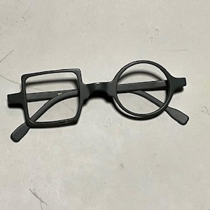 Może przedstawiać: Czarne okulary o unikalnym designie. Jedna soczewka jest kwadratowa, druga okrągła. Oprawki i zauszniki są matowe czarne. Okulary są na jasnoszarym tle.