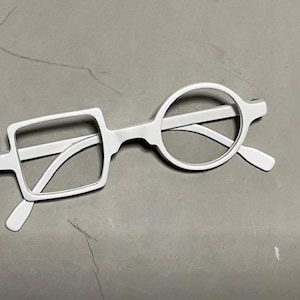 Może przedstawiać: Białe okulary o unikalnym designie. Jedna soczewka jest kwadratowa, druga okrągła. Oprawki i zauszniki są białe. Okulary są na szarym tle.