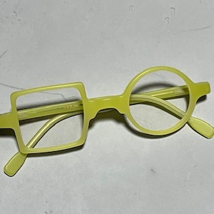 Może przedstawiać: Unikalne, limonkowo-zielone okulary z kwadratową soczewką po lewej stronie i okrągłą soczewką po prawej stronie. Oprawki mają charakterystyczny, nowoczesny design. Okulary są na neutralnym tle.
