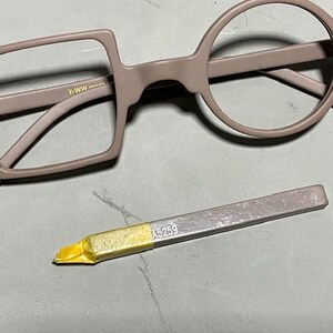 May include: A pair of eyeglasses with a unique design, featuring one square and one round lens frame in a muted mauve color. A small, silver-colored tool with the number "A5259" is also present.