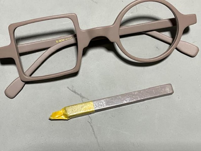 May include: A pair of eyeglasses with a unique design, featuring one square and one round lens frame in a muted mauve color. A small, silver-colored tool with the number "A5259" is also present.