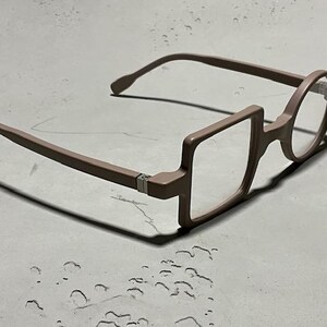 May include: Unique eyeglasses with a brown frame. The design features a combination of square and round lenses. The glasses are set against a neutral background, highlighting their distinctive shape and style. A modern eyewear accessory.