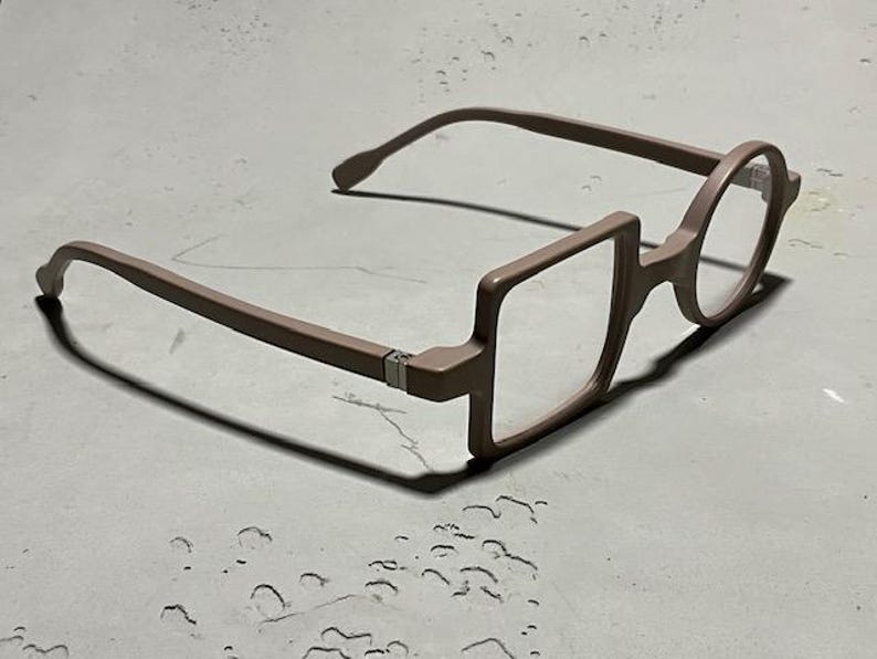 May include: Unique eyeglasses with a brown frame. The design features a combination of square and round lenses. The glasses are set against a neutral background, highlighting their distinctive shape and style. A modern eyewear accessory.