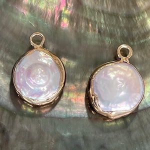 May include: Three irregularly shaped, white pearl pendants with gold-colored metal edging. Each pendant has a small loop at the top for hanging. The pearls have a lustrous, iridescent sheen, and are set against a dark, textured background.
