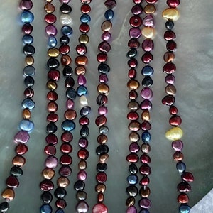 May include: Several strands of colorful freshwater pearl necklaces. The pearls are in shades of red, pink, blue, gold, and black. The necklaces are displayed on a large, iridescent, shell-shaped surface.