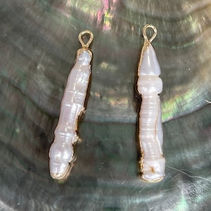 May include: Two white pearl pendants with gold accents. The pearls are shaped like a long, thin, pointed teardrop. The pendants are hanging from a gold loop.